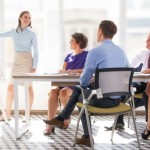 Cheerful young businesswoman pointing at whiteboard and explaining strategy. Confident business coach presenting project to staff. Colleagues listening to presenter. Business meeting concept